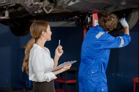 Mechaniker überprüft Auto von unten, während eine Frau mit Klemmbrett Notizen macht