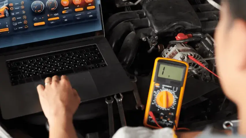Mechaniker misst Spannung mit Multimeter am Motor, während Fahrzeugdiagnose auf Laptop läuft